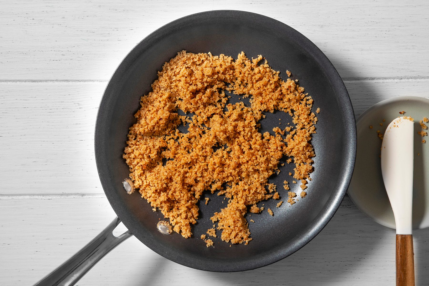 A non-stick frying pan contains golden-brown toasted breadcrumbs. To the right, there's a small white spatula resting on a white dish, all set on a white wooden surface.