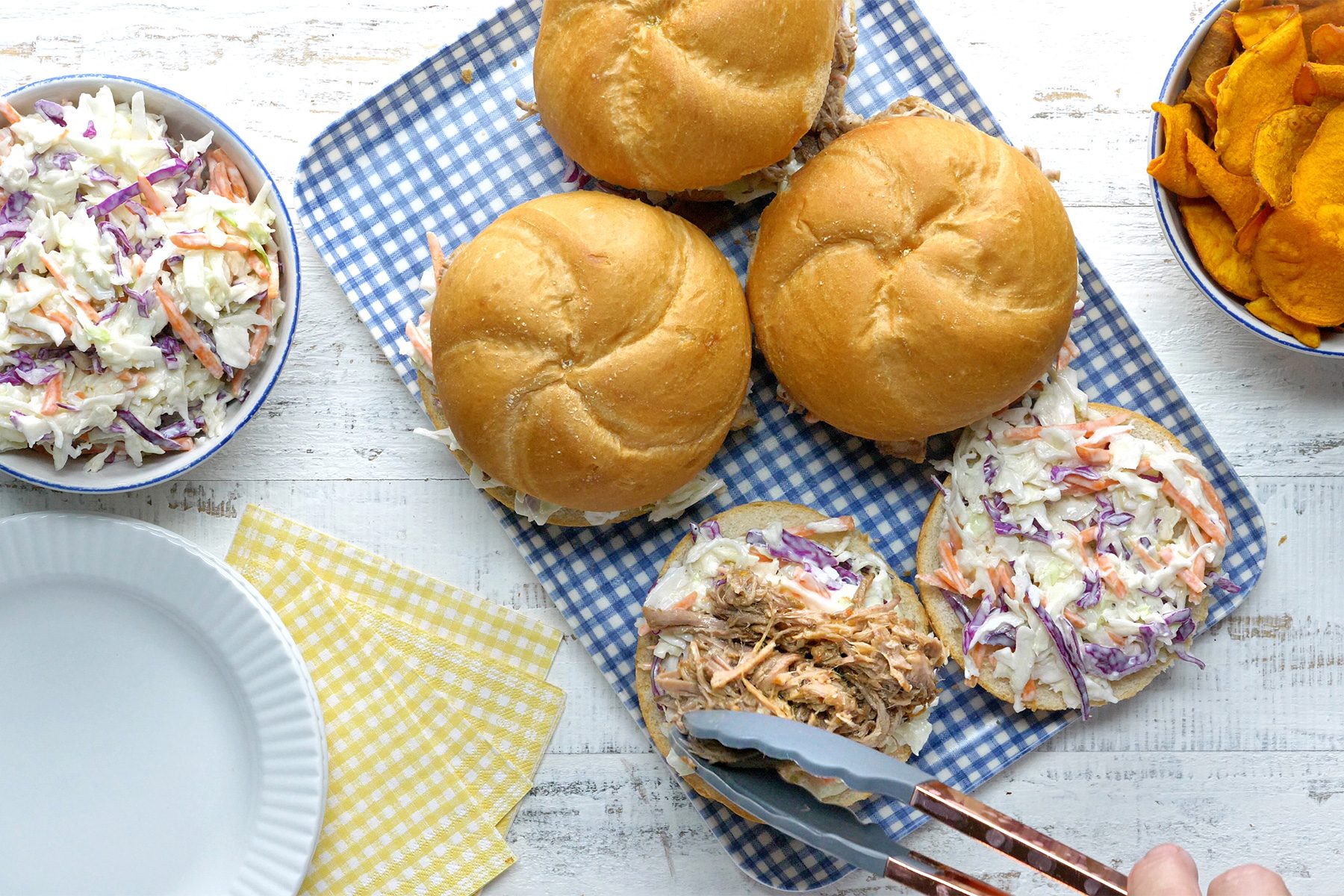 overhead shot; white textured background; Carolina-Style Pork Barbecue served over a tray with salad in small bowl, a small white over kitchen napkin; adding pork mixture over bun;