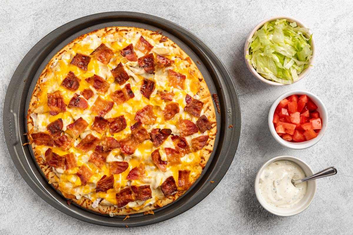 Taste of Home Chicken Bacon Ranch Pizza recipe photo of the pizza fully baked with sides of tomato, lettuce, and ranch dressing for toppings.