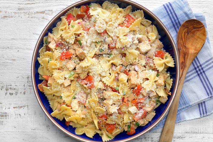 A bowl of bow-tie pasta mixed with chunks of chicken, red bell peppers, and green herbs, topped with grated Parmesan cheese.