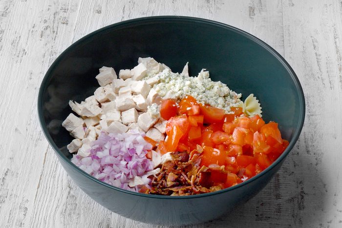 A black bowl on a white wooden surface contains a mixture of cubed chicken, blue cheese crumbles, diced tomatoes, finely chopped red onions, and crumbled bacon.