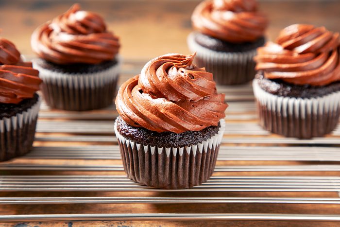 Close-up of five chocolate cupcakes topped with swirls of chocolate frosting, placed on a cooling rack. The cupcakes are displayed in a well-lit setting, emphasizing the rich texture of the frosting and the moist appearance of the cupcakes.