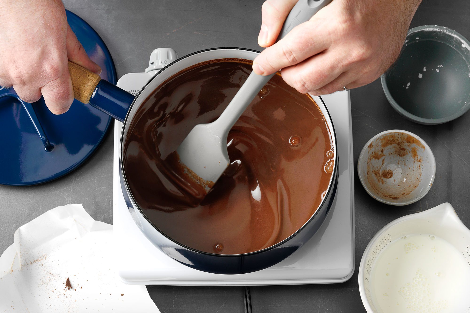 A person stirs melted chocolate in a blue pot with a spatula on a stove; marble surface;