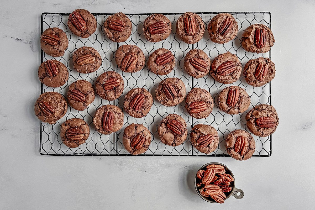 Taste of Home German chocolate cookies on a wire cooling rack with a tin cup of pecans on a marble surface.