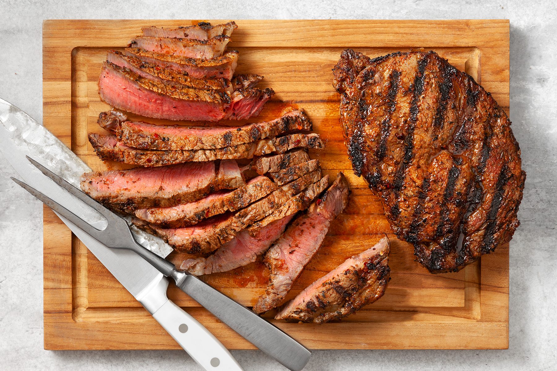 A wooden cutting board with a grilled steak, partially sliced. The steak has visible grill marks and is medium-rare in the center.
