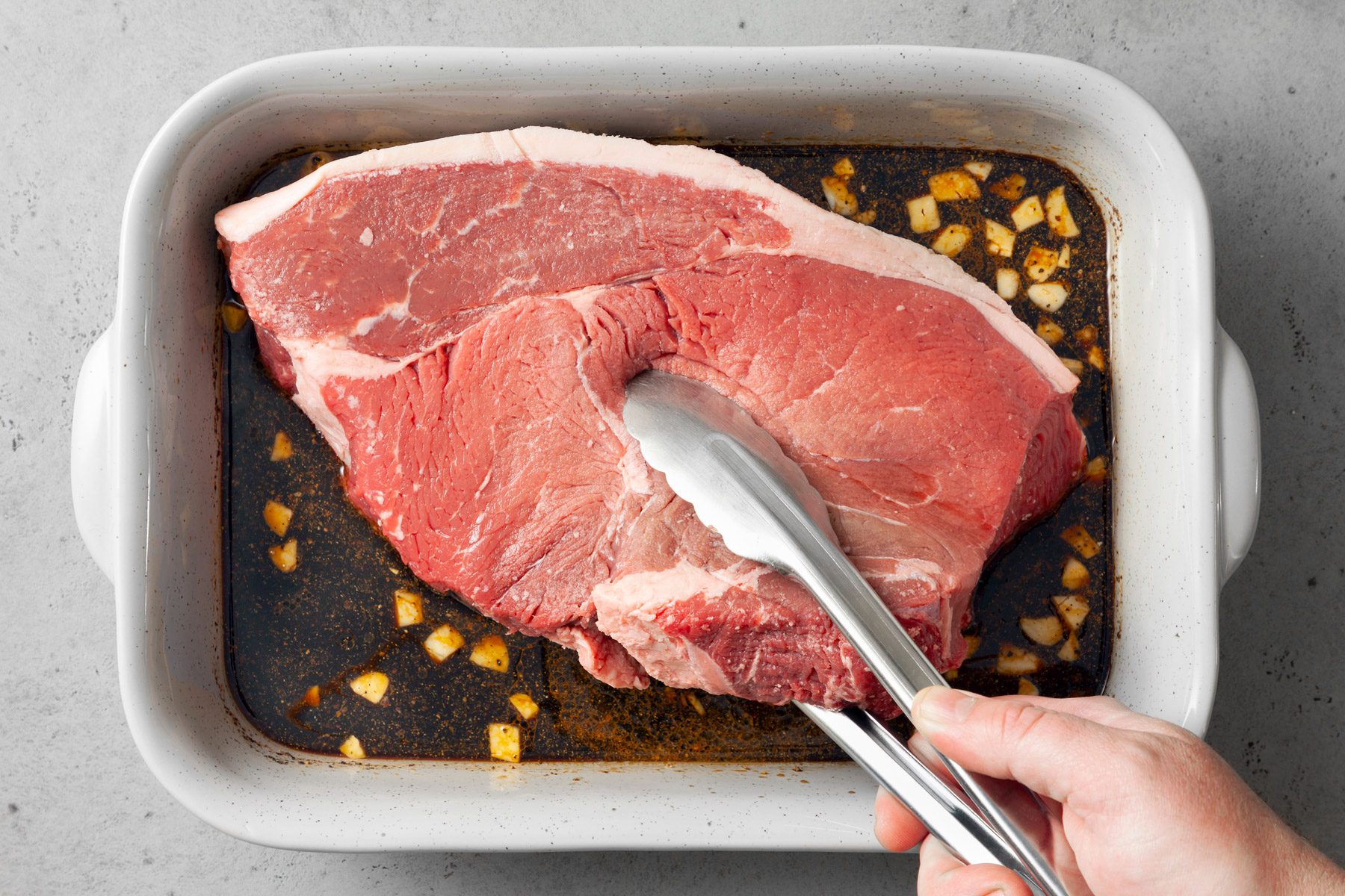 A large piece of raw beef is being placed in a marinade consisting of chopped garlic and liquid in a white baking dish. A person is holding the beef with metal tongs over the dish.
