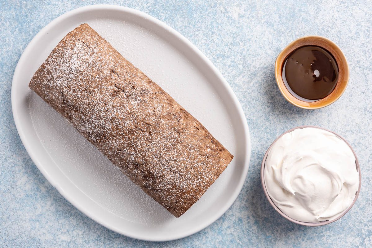 Taste of Home Ice Cream Cake Roll recipe photo the cake rolled up sprinkled with powdered sugar and sides of chocolate sauce and whipped cream.