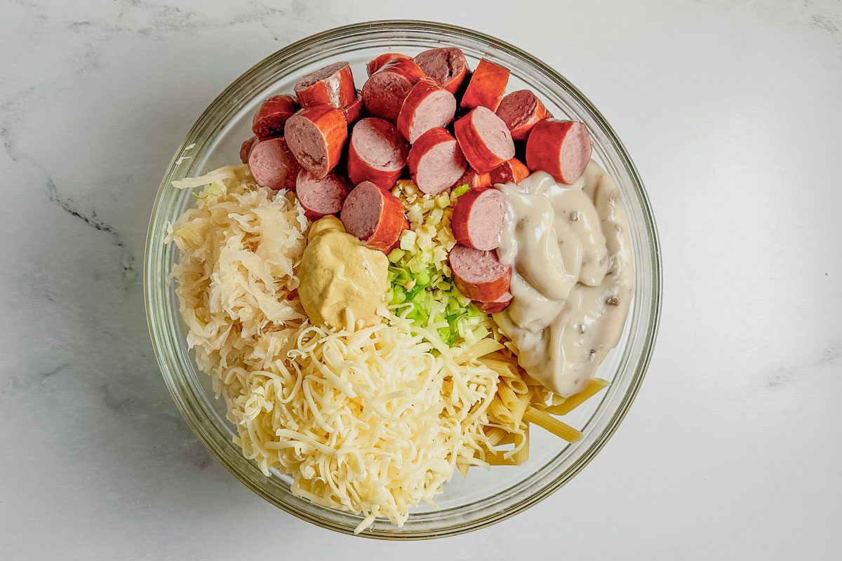 Kielbasa, cheese, sauerkraut, mustard, scallions and cream of mushroom soup in a glass bowl on a marble surface for Taste of Home kielbasa casserole.