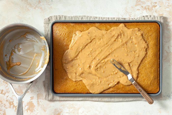 overhead shot; white textured background; Spooning glaze mixture over warm cake, spreading evenly with spreader;
