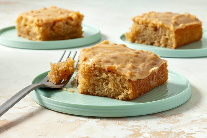 close shot; white textured background; Peanut Butter Sheet Cake cut into square pieces served on three small plates with silver fork;