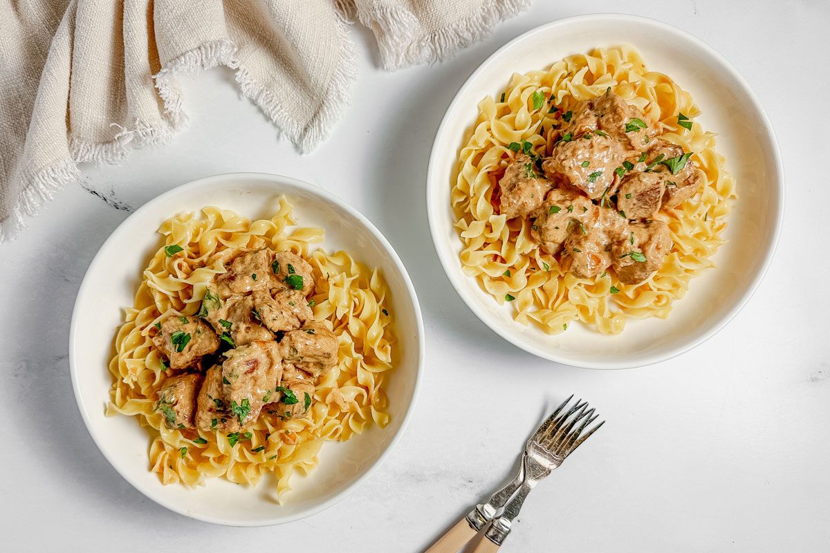 Taste of Home pork stroganoff in white ceramic bowls with a cloth napkin on a marble surface.