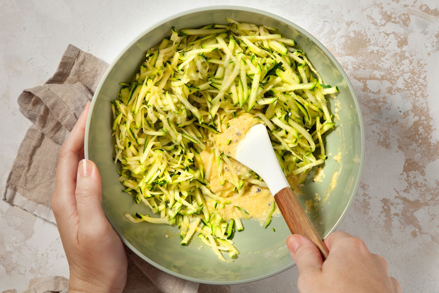 Hands mix shredded zucchini into batter in a bowl using a white spatula with a wooden handle. A light brown cloth is partially visible next to the bowl on a beige, textured surface.
