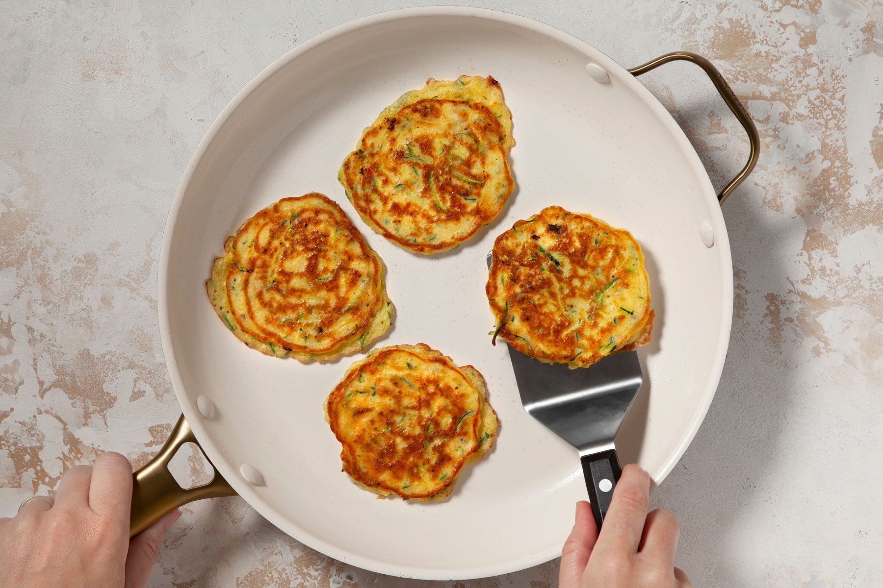 A person is cooking four zucchini fritters in a skillet. The person is using a spatula to lift one of the fritters. The background surface is light and textured.