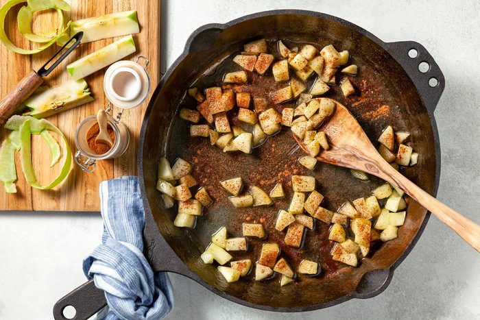 overhead shot; white background; a cast-iron skillet filled with diced apples, being sautéed in butter; The apples are a light brown color and are coated in a mixture of spices, including cinnamon and sugar; There is a wooden cutting board with apple peels and a small bowl of cinnamon on the side;