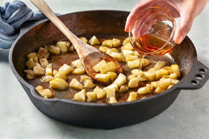 3/4th shot; white background; A cast-iron skillet with wooden spatula contains sautéed apples, coated in a mixture of spices and butter; A person's hand is pouring a liquid into the skillet;