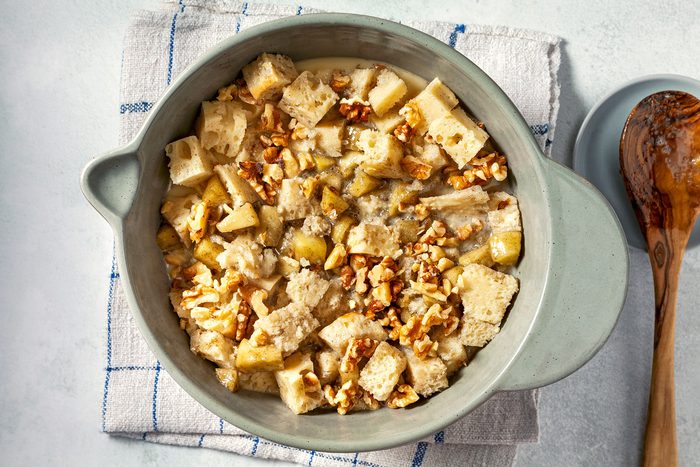 overhead shot; white background; A grey bowl over kitchen towel contains a mixture of bread cubes, apples, walnuts, and a creamy custard; The bread cubes are partially submerged in the custard, and the walnuts are scattered throughout the mixture; A wooden spoon rests on the rim of the bowl, a wooden spatula is placed over small plate;