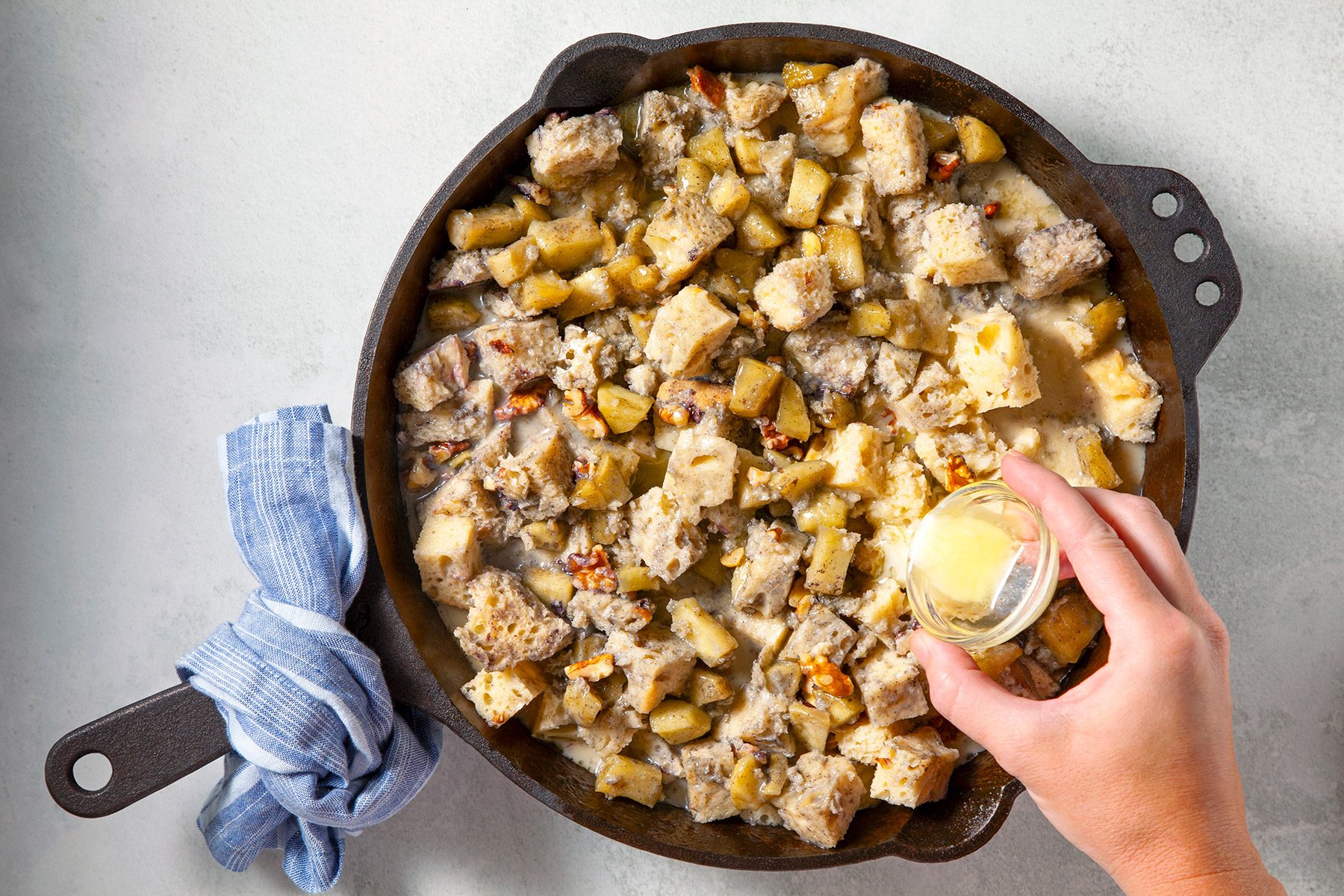 overhead shot; white background; A cast-iron skillet contains a mixture of bread cubes, apples, walnuts, and a creamy custard; A person's hand is pouring a small amount of melted butter into the skillet;