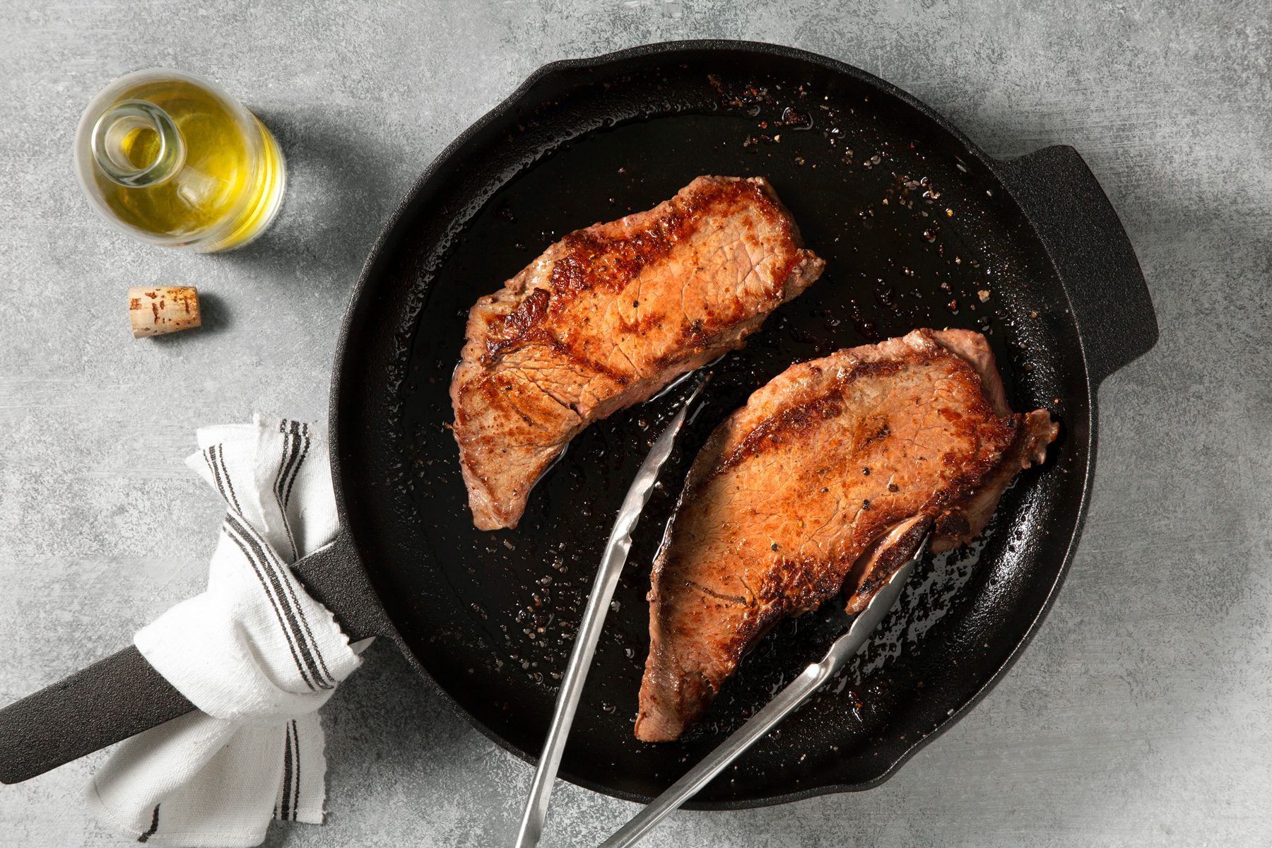 overhead shot; light grey background; in a large skillet, cooking steak using tongs, a bottle of oil is also seen nearby