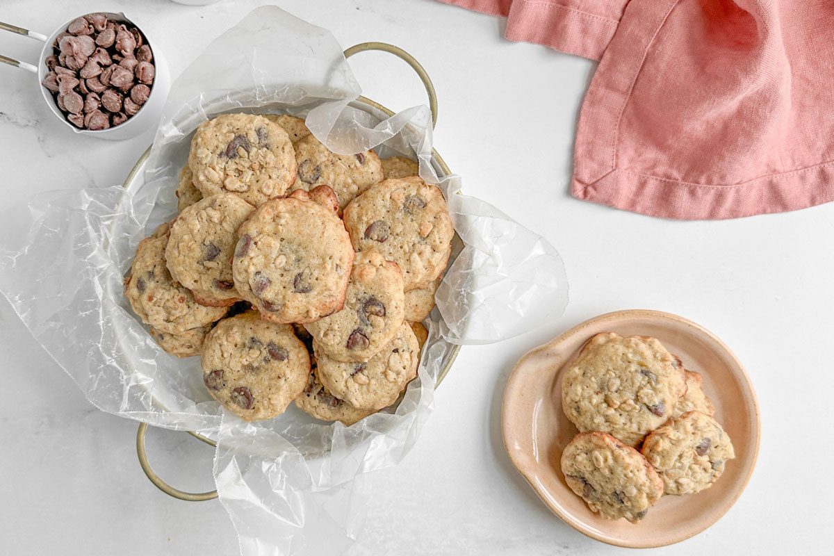 Taste of Home Banana Oatmeal Cookies with cups of oatmeal, chocolate chips and walnuts on a marble surface next to a pink linen napkin