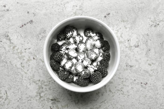 A white bowl filled with blackberries lightly dusted with powdered sugar sits on a textured, light gray surface. The contrast of dark berries and white sugar creates a visually striking effect.
