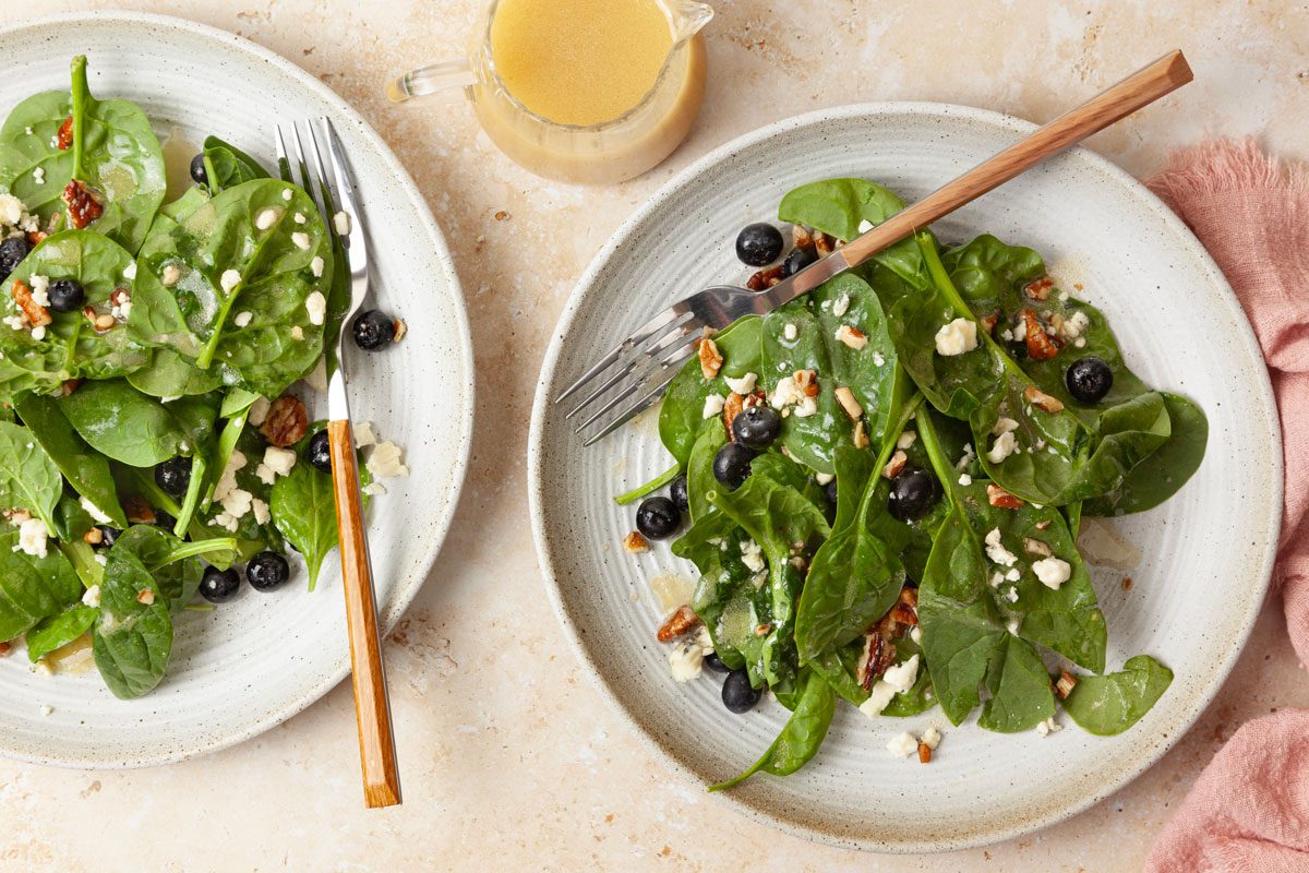 Two single servings of Taste of Home Blueberry Salad on light gray ceramic plates, wooden handled forks, pink linen, dressing cruet