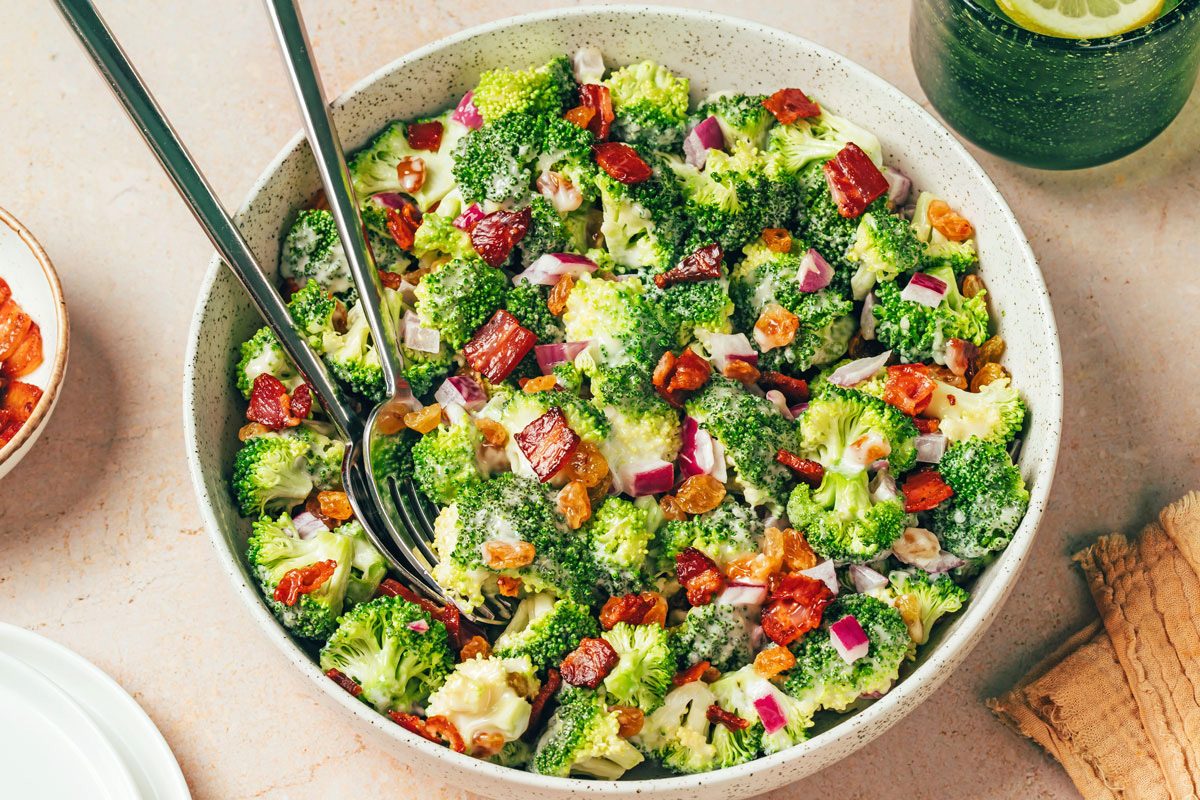 Broccoli Raisin Salad in a serving bowl with serving spoon and topped with cooked ham, recipe for Taste of Home