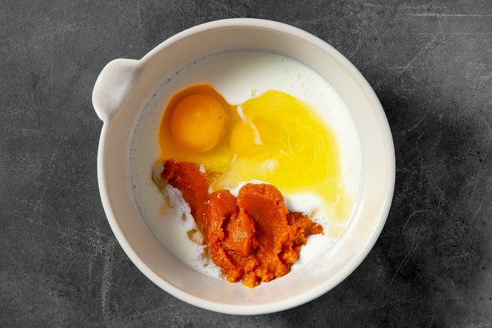 A white bowl containing an uncooked egg, a broken egg yolk, some milk, and a dollop of pumpkin puree, set against a dark, textured background.