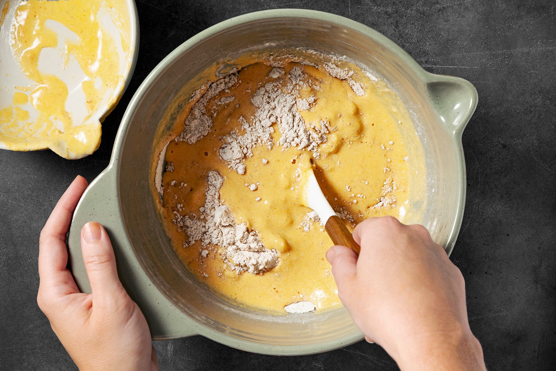 A person is mixing a yellow batter with a spatula in a large green bowl. The batter has some dry flour on top. To the left, a smaller bowl with some remaining batter is visible.