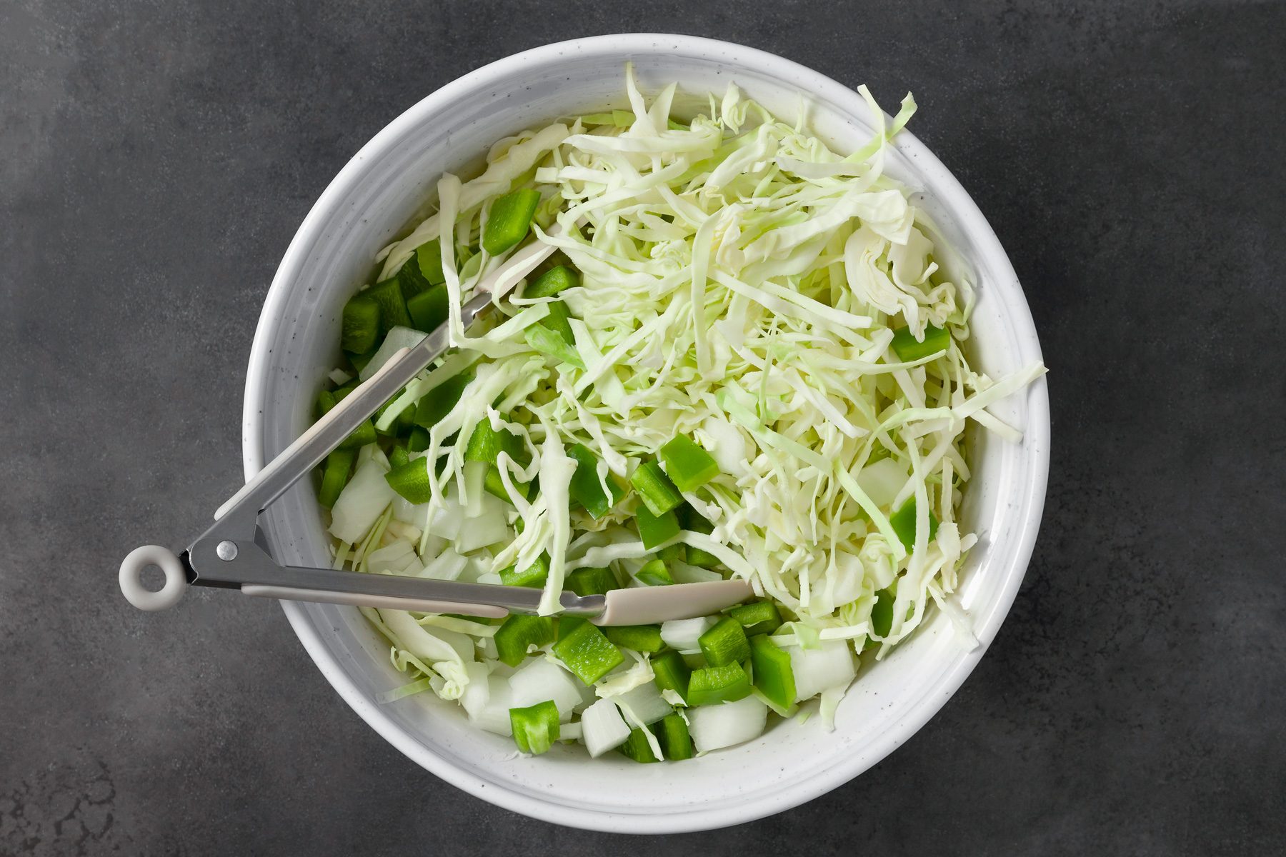 overhead shot; dark grey background; In a large bowl, combined cabbage, green pepper and onion; tongs is placed over bowl