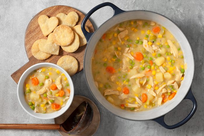overhead shot; light grey background; Chicken Potpie Soup served in a large, blue Dutch oven; A smaller bowl of the soup is also shown in the image; A variety of heart-shaped biscuits are arranged on a wooden cutting board; serving spoon is also placed over small wooden coaster