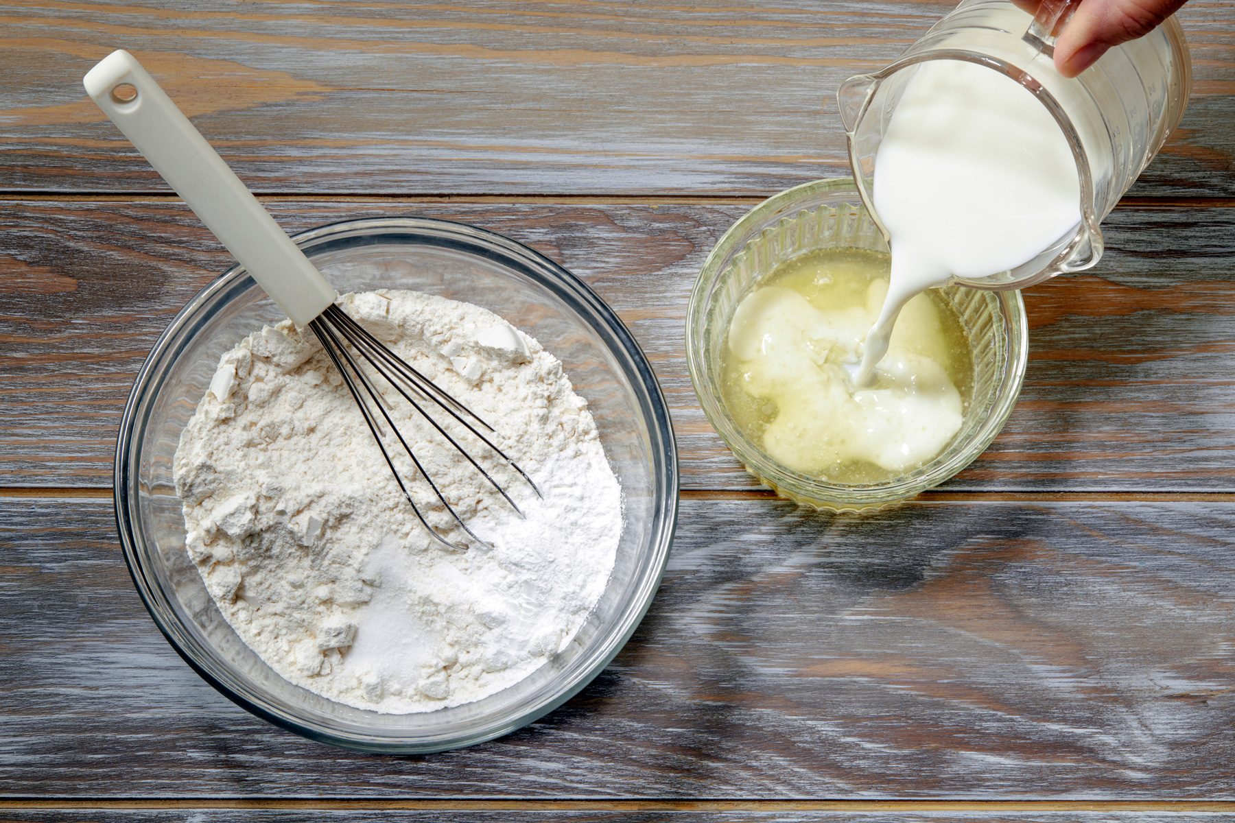 overhead shot; wooden background; flour, baking powder and salt in a bowl with whisker; pouring milk in melted butter