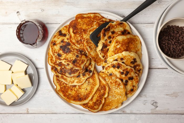 Full yield of Taste of Home Chocolate Chip Pancakes on a white platter, plate for butter pats, syrup cruet, mini chocolate chips