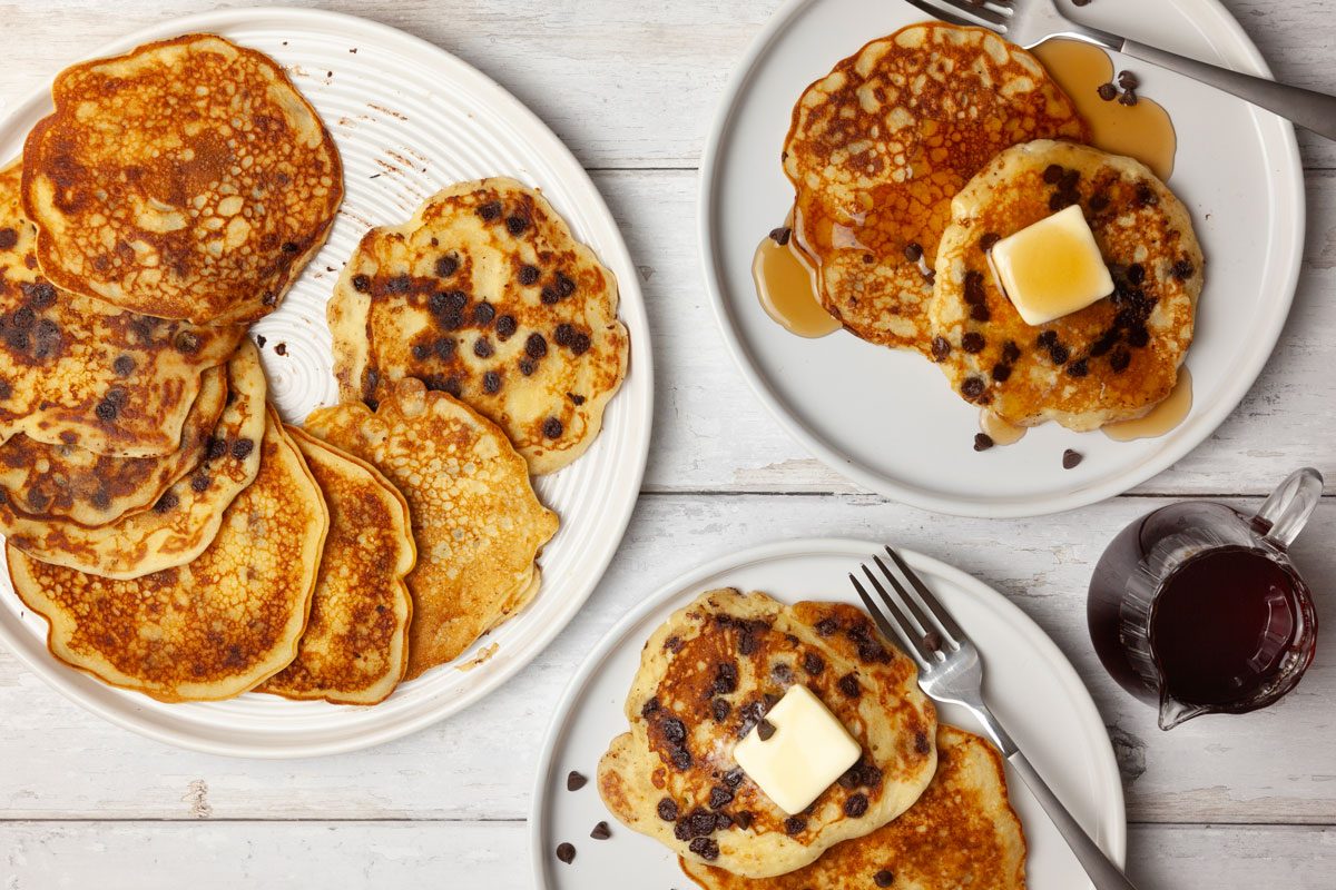 Full yield of Taste of Home Chocolate Chip Pancakes on a white platter, two single servings, plate for butter pats, syrup cruet, mini chocolate chips