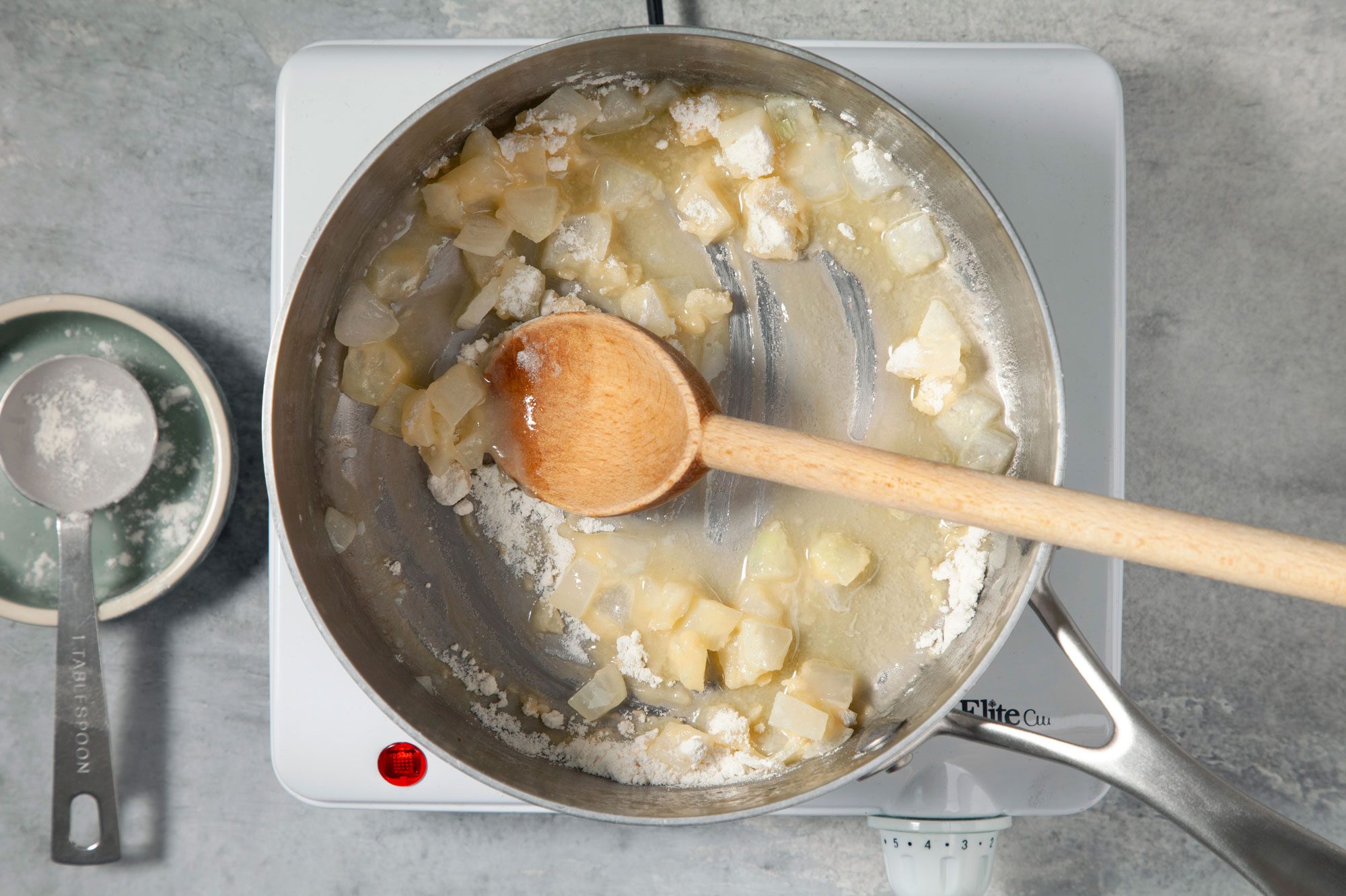 A wooden spoon is stirring the mixture, A measuring cup filled with flour sits on the countertop next to the saucepan