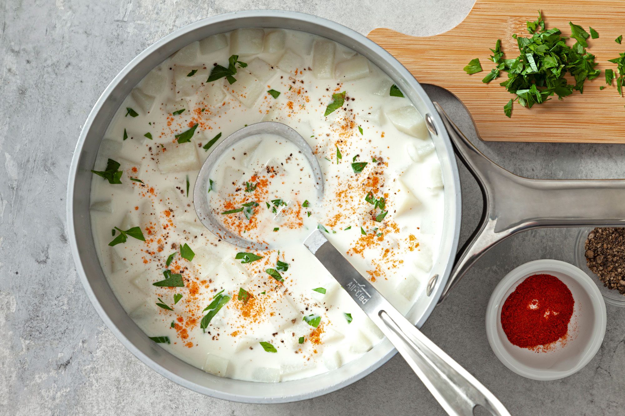Cream of Potato Soup in a large saucepan with large serving spoon in it and paprika powder in small bowl and minced fresh parsley on wooden board