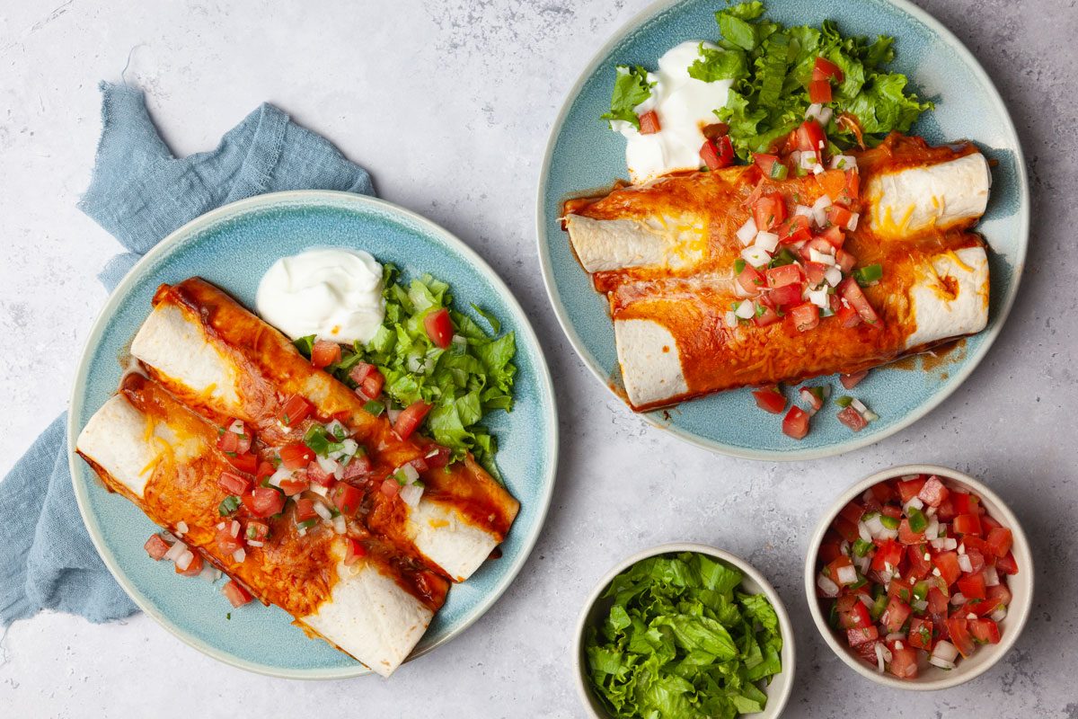 Two single servings of Taste of Home Crock-Pot Chicken Enchiladas garnished with pice de gallo, lettuce and sour cream. Small bowls of pico and lettuce.