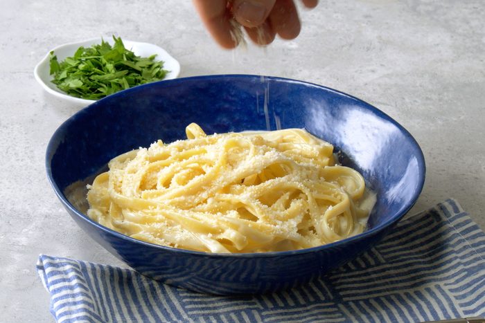 Sprinkling with Parmigiano Reggiano over the Fettuccine Alfredo served in a large blue bowl over kitchen towe