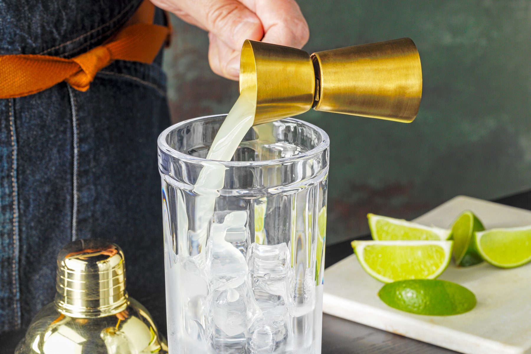 High angle shot of fill a shaker three-fourths full with ice; adding the gin and lime juice; shaker lid; chopping board; lime wedges; green and brown texture wall; black wooden base