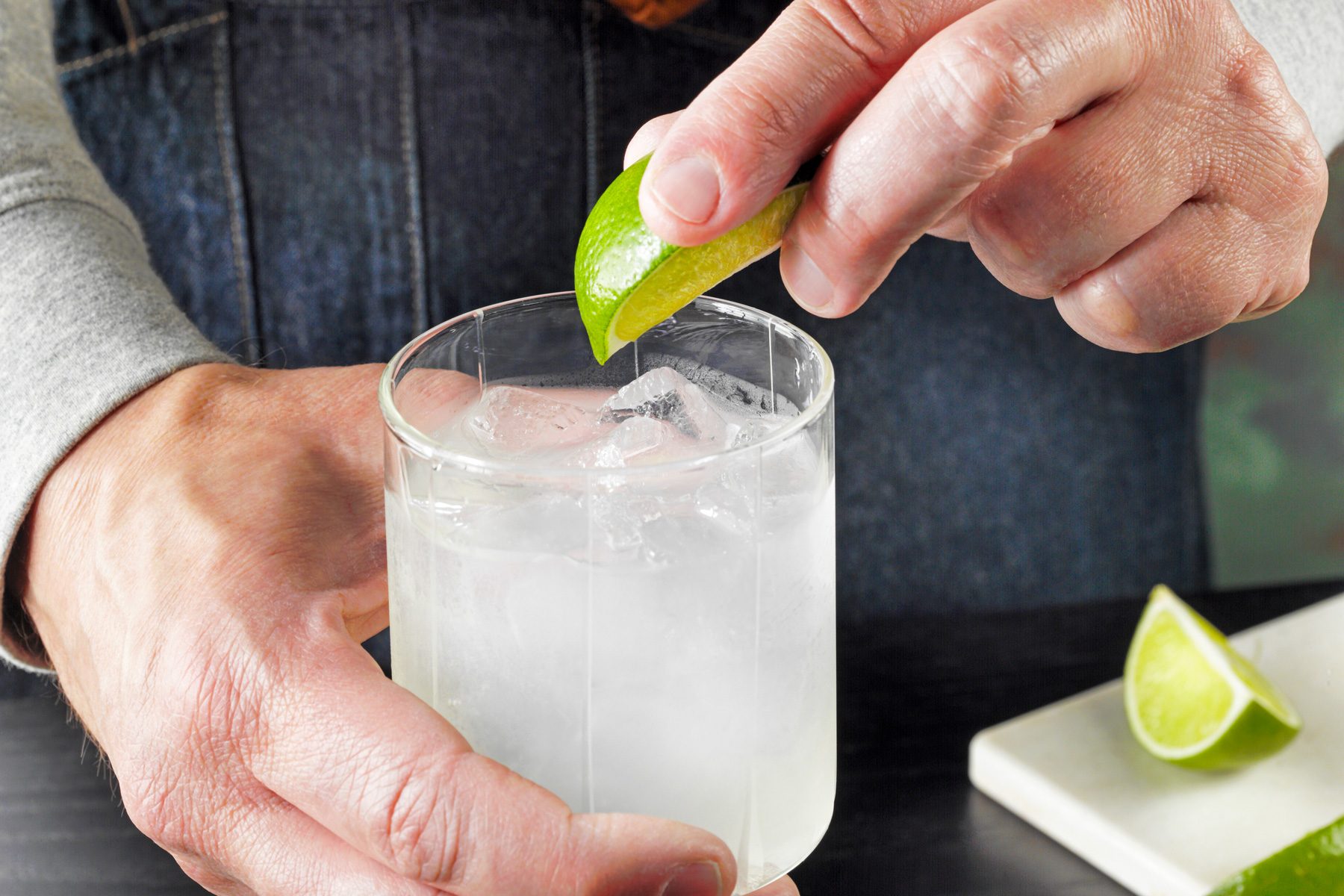 3/4 angle view shot of Gimlet; garnishing with lime; lime wedges; chopping board; green and brown texture wall; black wooden base