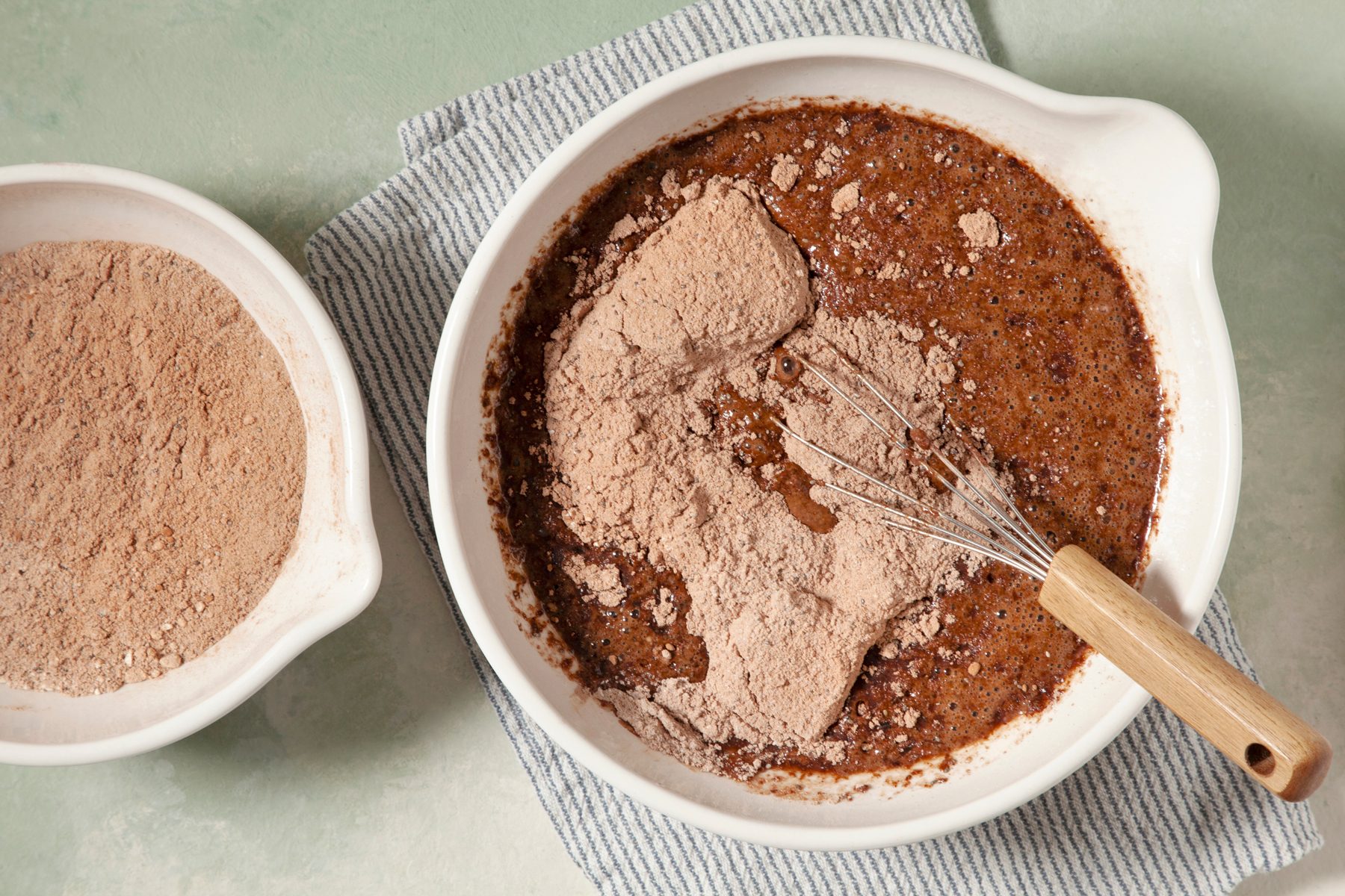 overhead shot; light green textured background; A white bowl contains a dark brown mixture of ingredients; a whisker rest inside the bowl placed over kitchen towel; Dry ingredients, such as cocoa powder and flour, are visible on the surface of the batter; Another white bowl filled with dry ingredients is in the background