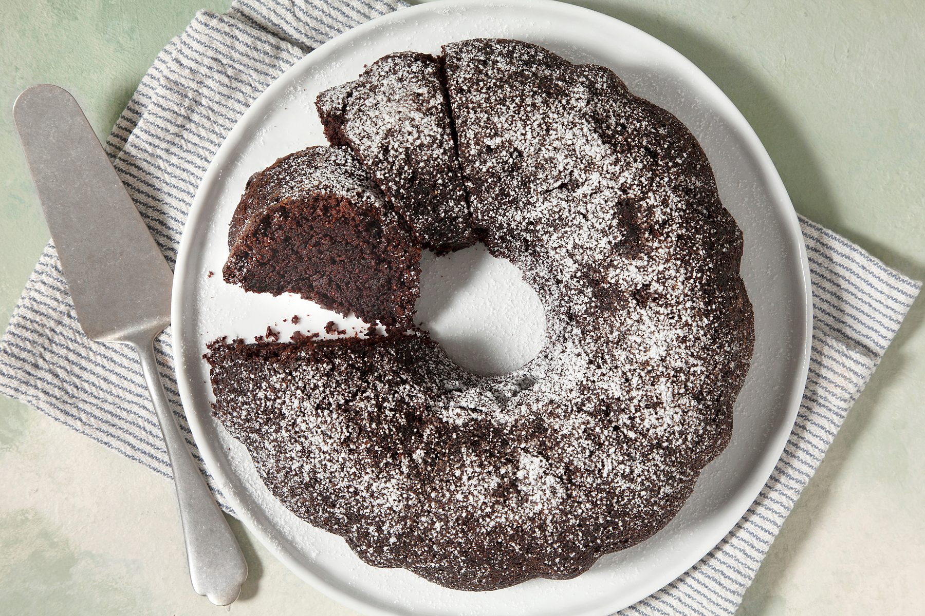 overhead shot; light green textured background; A chocolate cake is displayed on a white plate; It's dusted with powdered sugar and appears to be a bundt cake with a central hole
