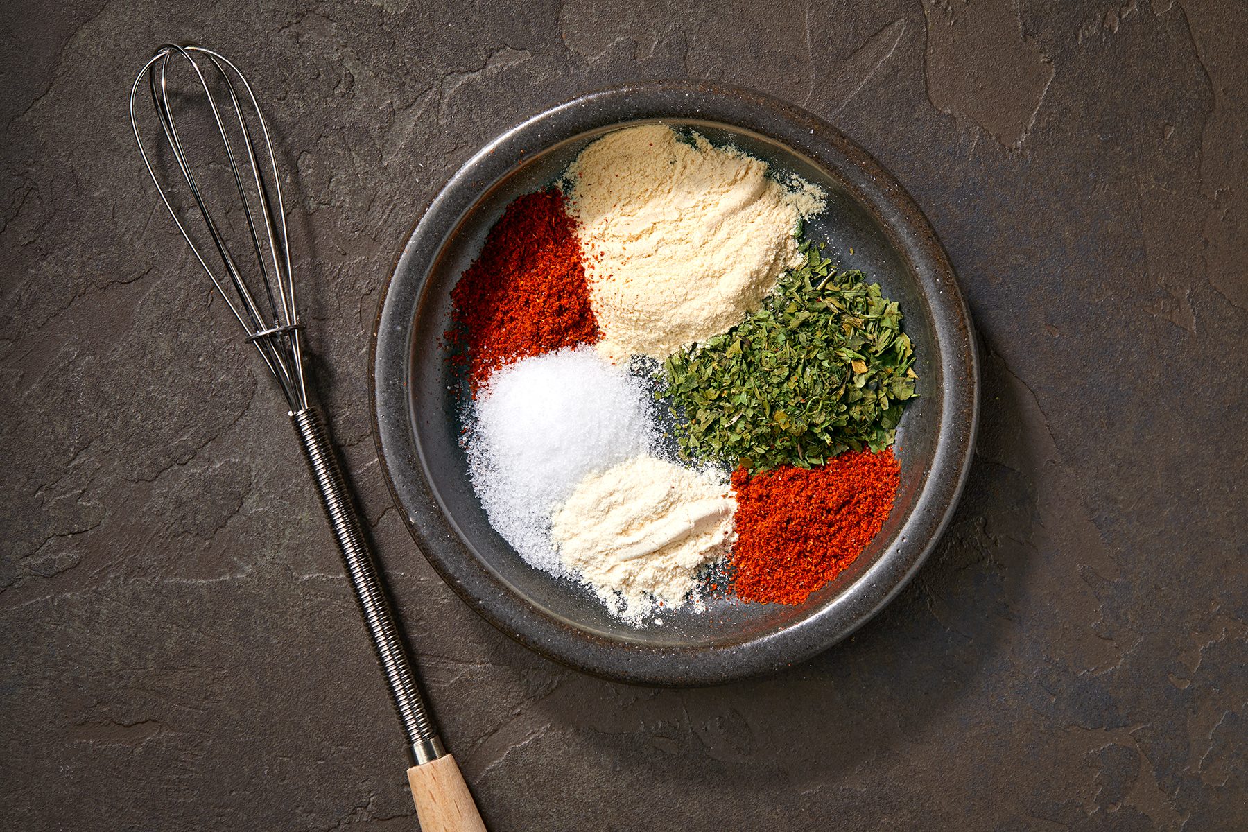 A ceramic bowl contains five types of seasonings arranged in separate mounds: dried herbs, paprika, salt, onion powder, and garlic powder. The bowl is placed on a dark, textured surface next to a small whisk with a wooden handle.