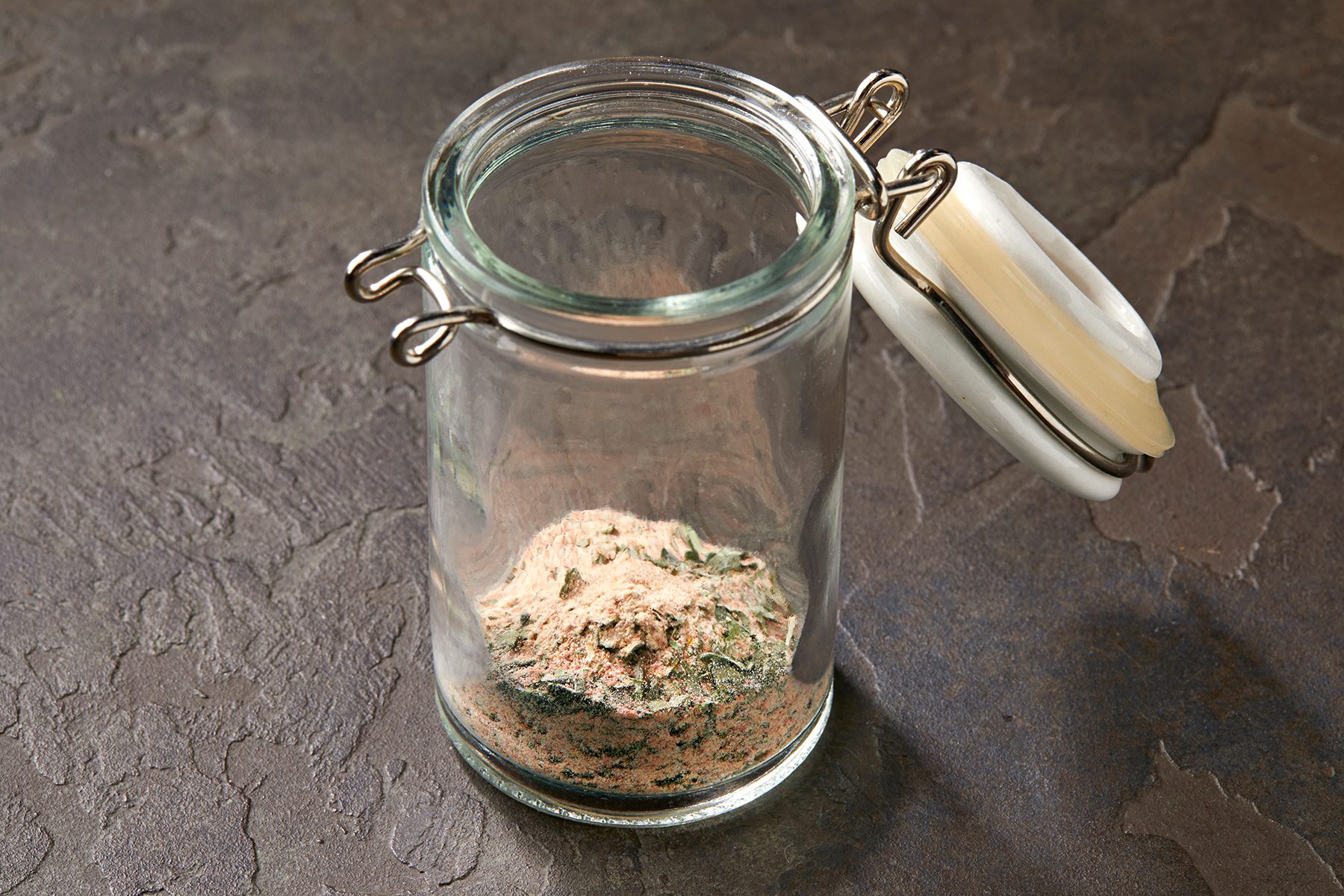 A clear glass jar with a hinged white lid contains a small amount of mixed powdered substance in various shades of brown, beige, and green. The jar stands on a textured, dark-colored surface.