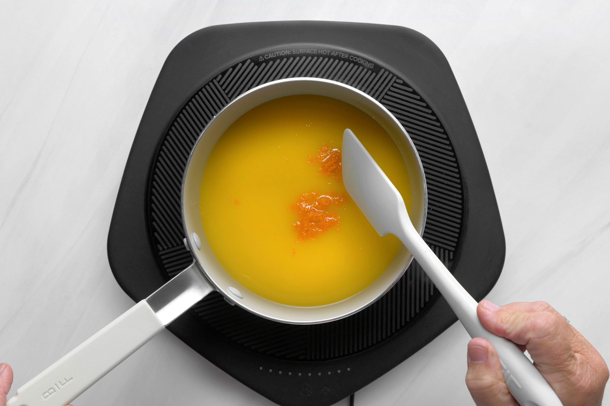 overhead shot; white background; A person is stirring a saucepan of liquid with a spatula, The liquid is a vibrant orange color and has small pieces of orange zest floating in it, The saucepan is placed on an electric induction cooktop;