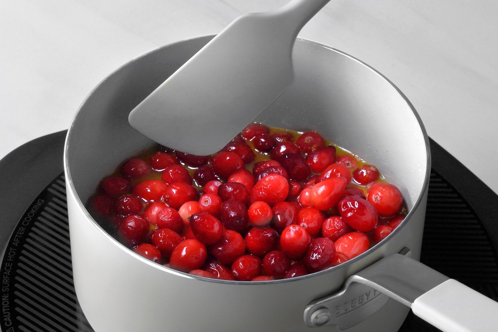 3/4th shot; white background; A saucepan on an electric induction cooktop contains a mixture of cranberries and a liquid, likely orange juice or sugar syrup, The cranberries are a vibrant red color and are simmering in the liquid, A spatula is being used to stir the mixture;