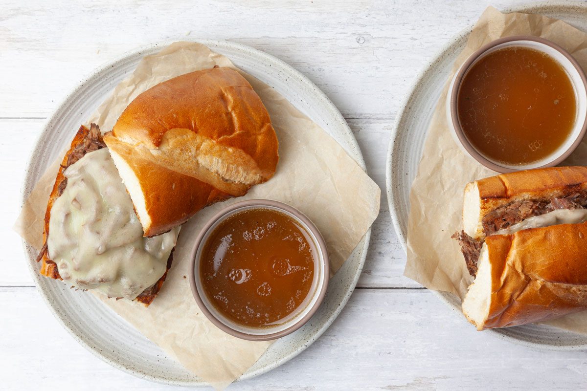 Two servings of Taste of Home Instant Pot French Dip with au jus on the side, provolone cheese, white wood surface
