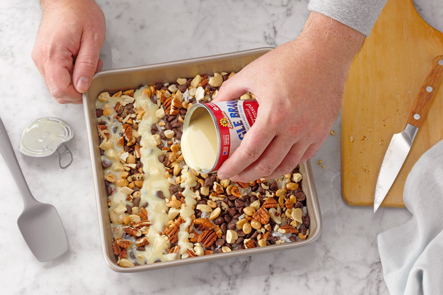 A person is pouring a can of sweetened condensed milk over a layer of mixed nuts, chocolate chips, and shredded coconut in a square baking pan. A spatula, measuring cup, and knife are on the marble countertop beside them.