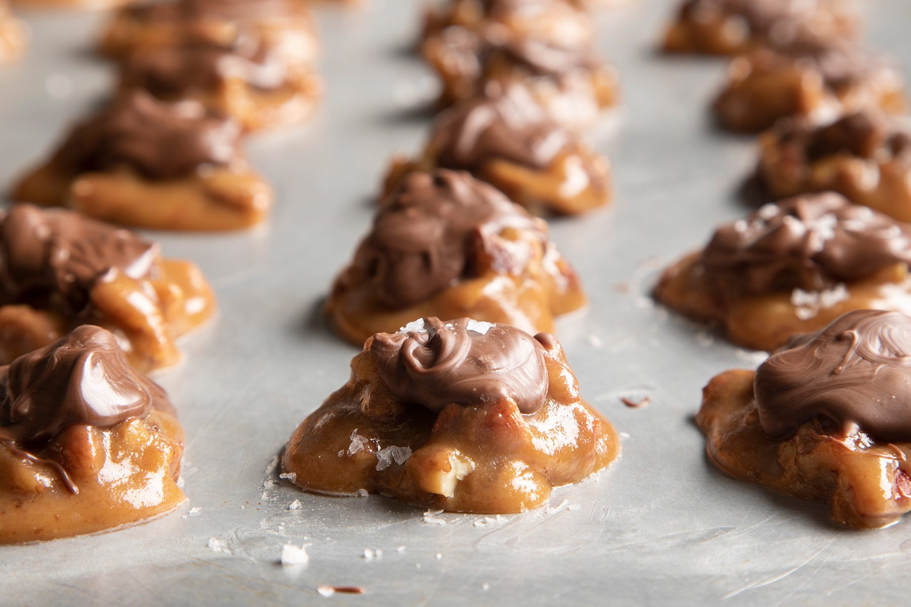 Close-up of multiple clusters of homemade chocolate and caramel pecan candies on a parchment-lined surface. The caramel is golden brown and topped with swirls of glossy melted chocolate. The candies are placed in rows, ready to set and cool.