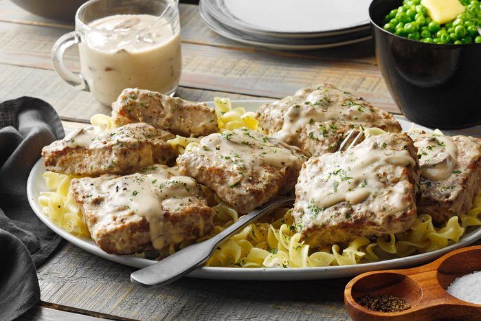 A platter of breaded meat topped with creamy white gravy, garnished with herbs, and served over a bed of egg noodles. A spoon rests on the platter. In the background, there are bowls of peas and sauce, along with a few empty plates.