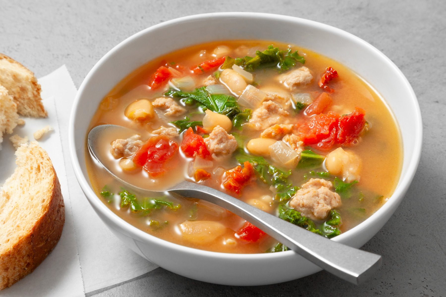 Table view shot of Italian Sausage Kale Soup; serve in a large bowl with bread; spoon; grey marble surface